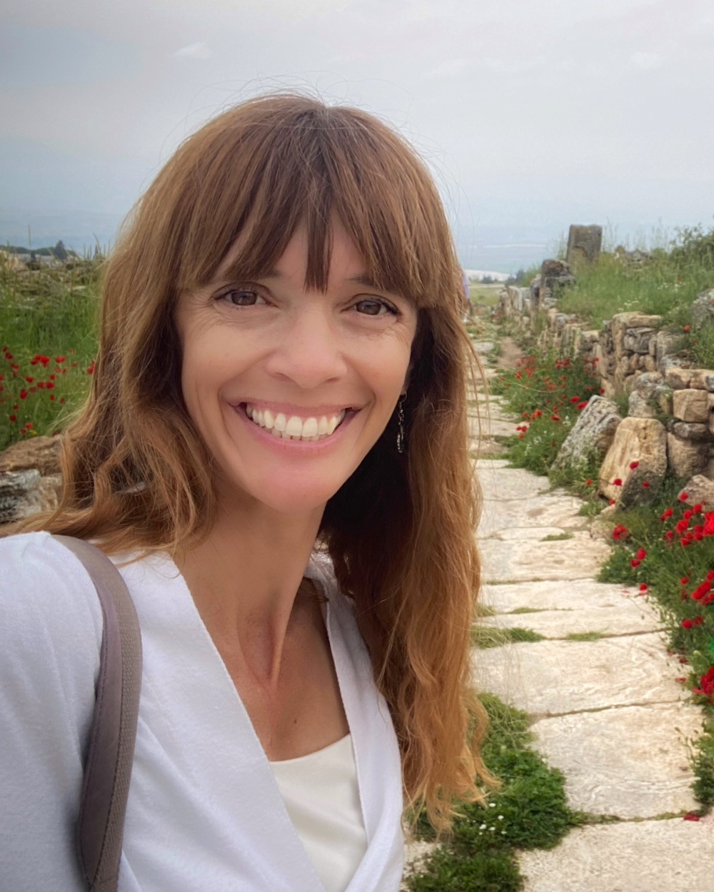 Daniela smiles wearing a white sweater in front of red flowers lining a stone path towards the ocean.