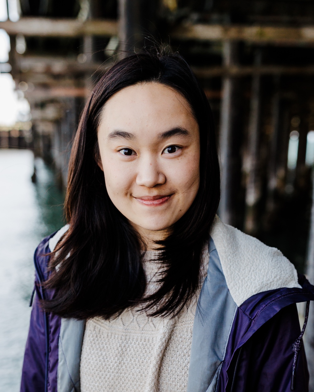 Esther smiles at the camera under a pier, in a light sweater and purple jacket.