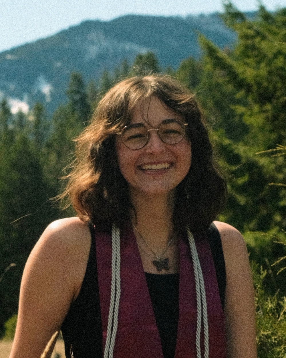 Katie smiles in front of a mountain and forest wearing a black dress and maroon stole.