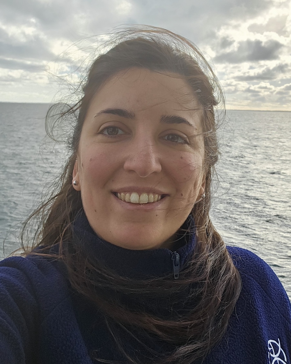 María smiles in front of ocean and sun-lit clouds.