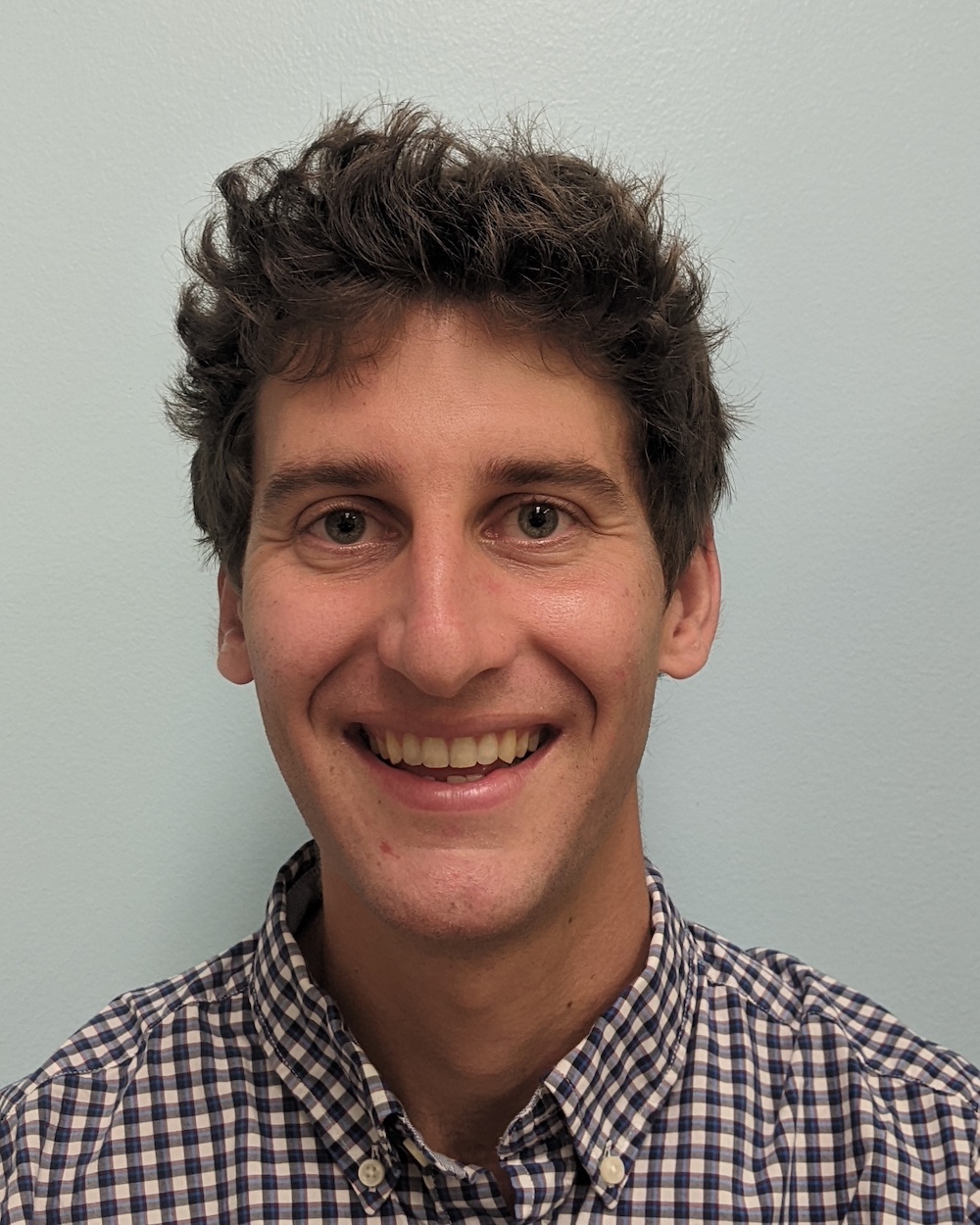 Nicholas smiles in a checkered shirt in front of a plain background.