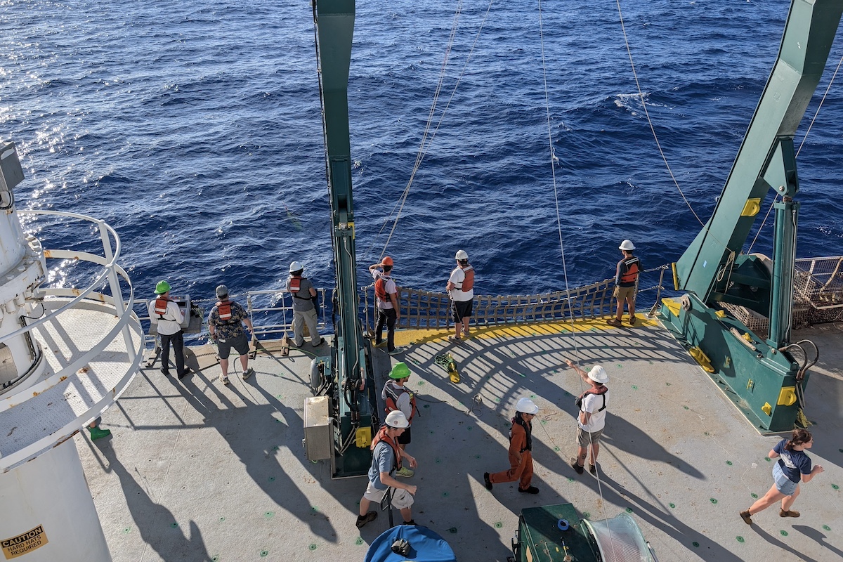 Scientists stand and walk on the deck of a research vessel. Some are walking while others stand by the edge of the ship and look out at the ocean.