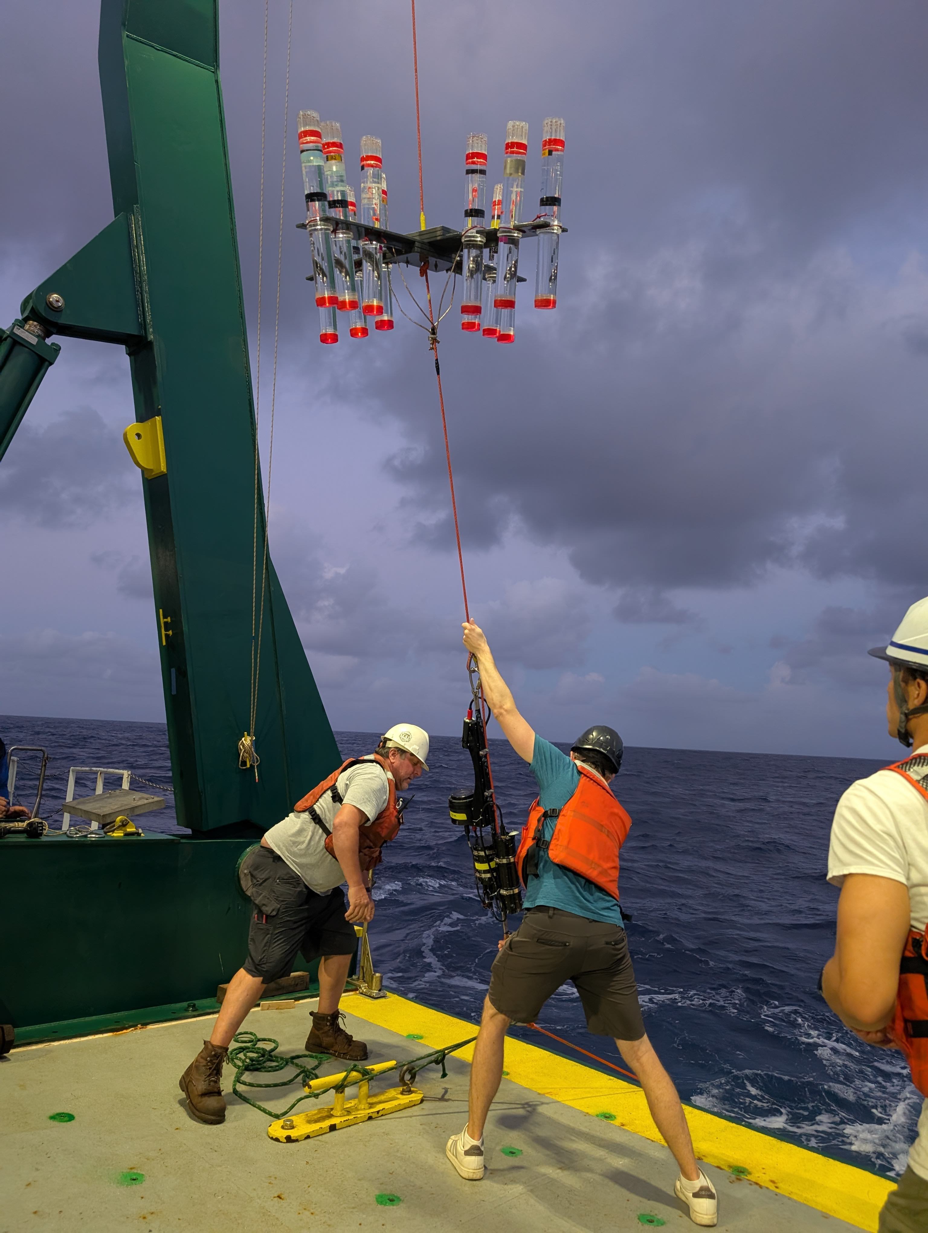 Two scientists wearing helmets and life vests heave a sediment trap array into the ocean at dusk.