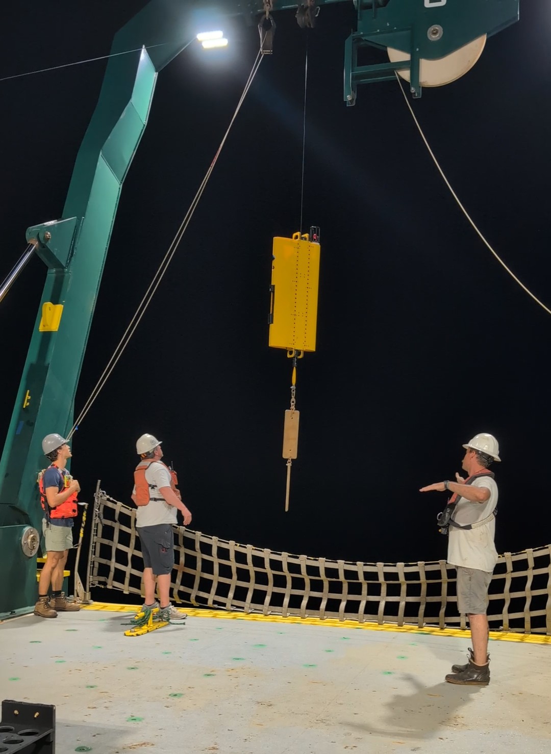 Scientists watch as a yellow apparatus hangs from a cable on a research vessel. The background is pitch black while the deck of the ship is well-lit.