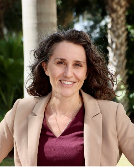 Hilary smiles at the camera, wearing a tan blazer and red shirt, in front of palms and greenery.