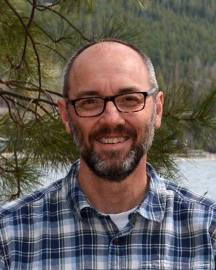 Matt smiles at the camera in front of pine needles, wearing a plaid shirt and black-framed glasses.