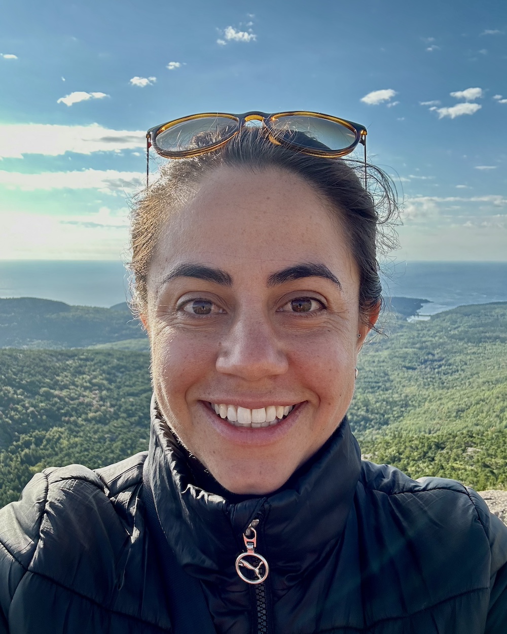 Raquel smiles in front of rolling green coastline and blue ocean in a black puffy jacket.