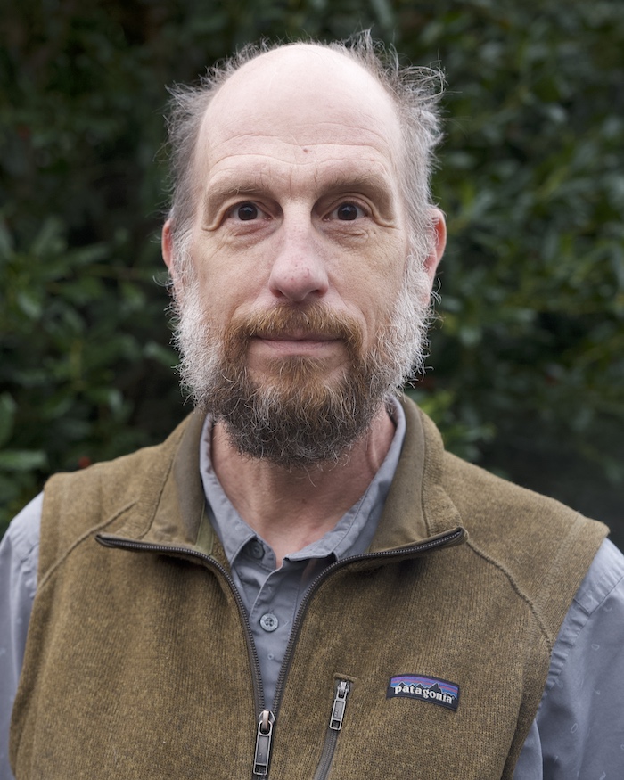 Bob looks at the camera in front of dark green leaves, wearing a button down and fleece vest.