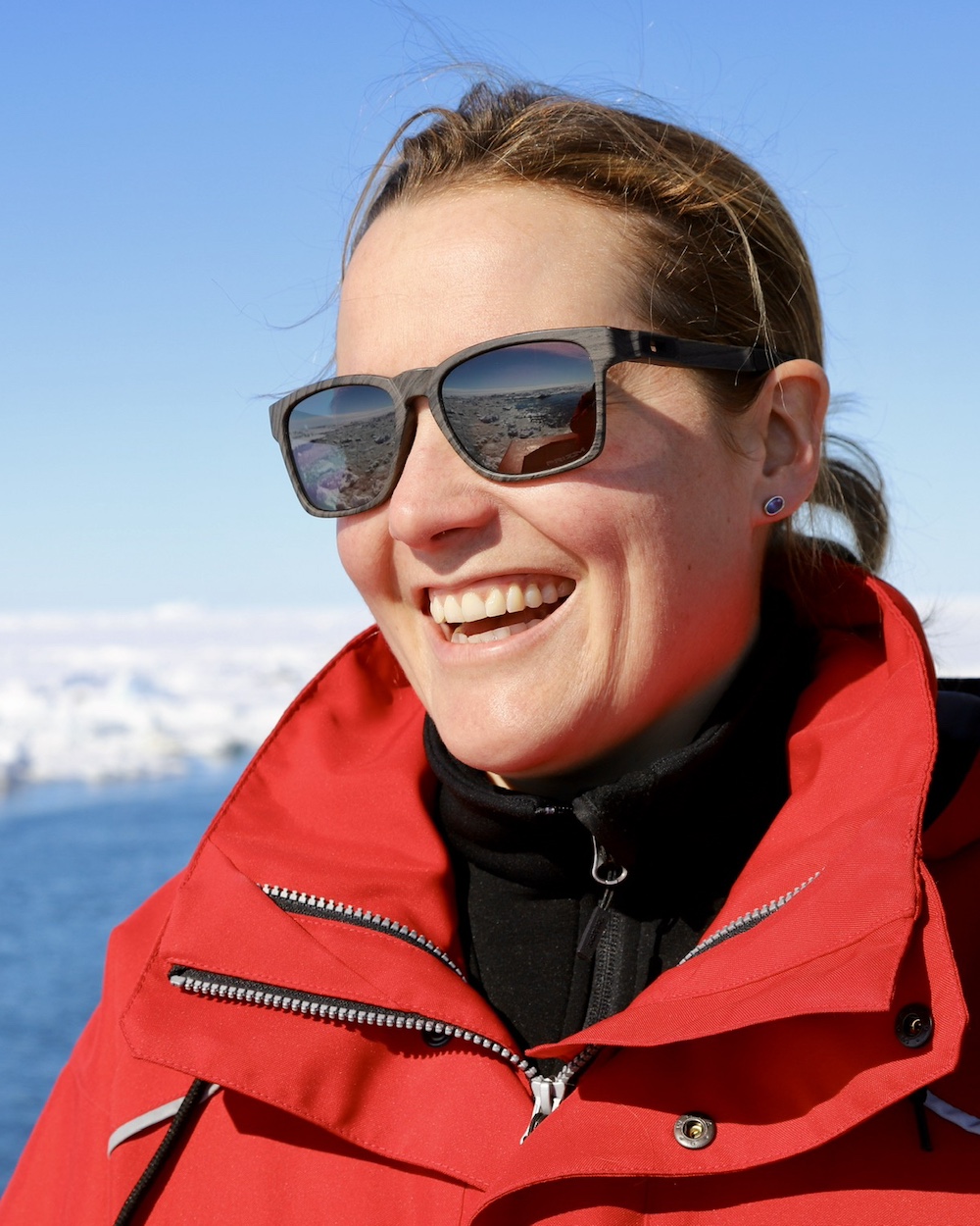 Sarah laughs, facing to the camera's left, in front of an expanse of sea ice and ocean.